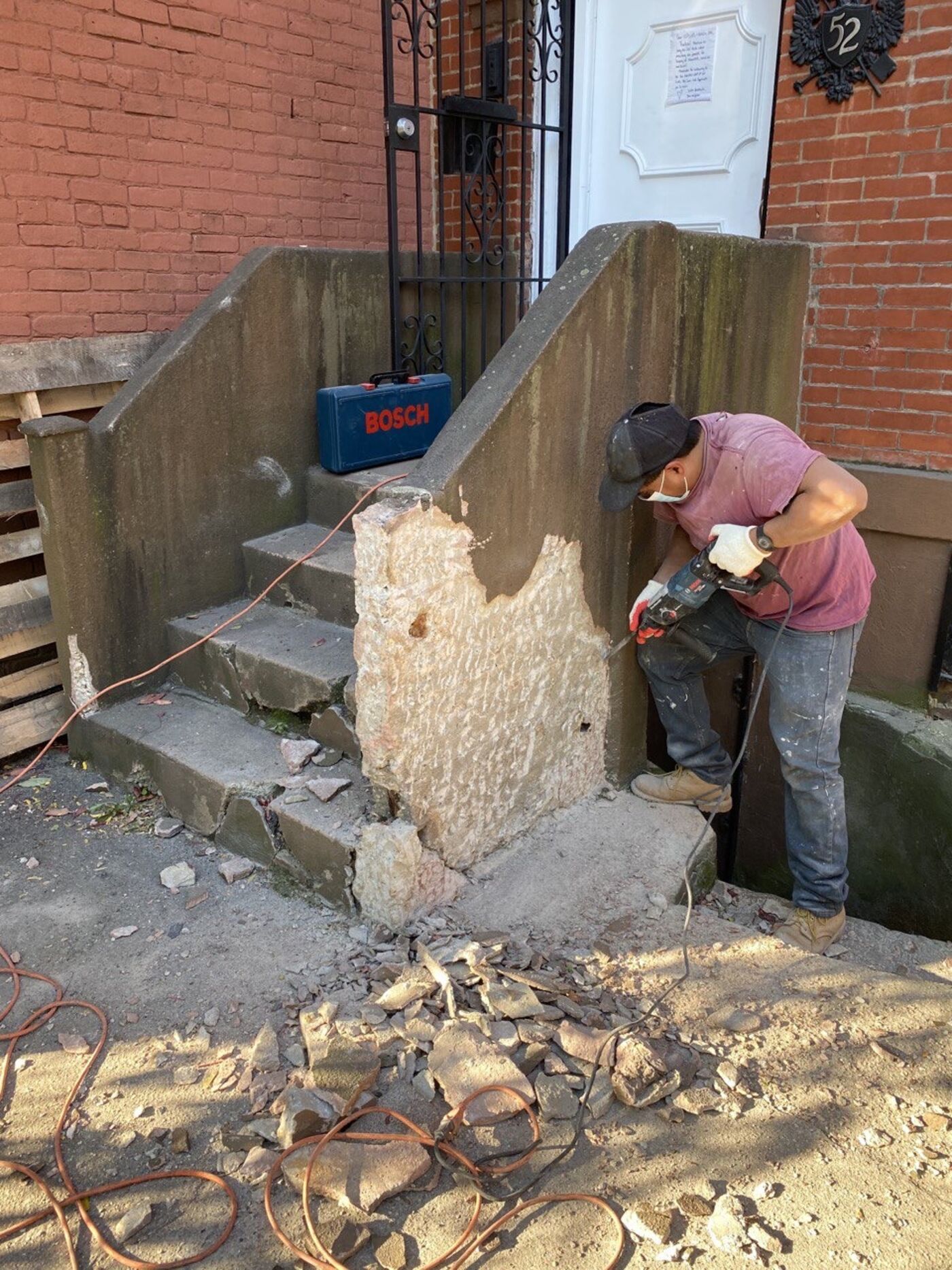 Worker using a power demolition hammer to remove damaged concrete from the front steps of a brick townhouse.